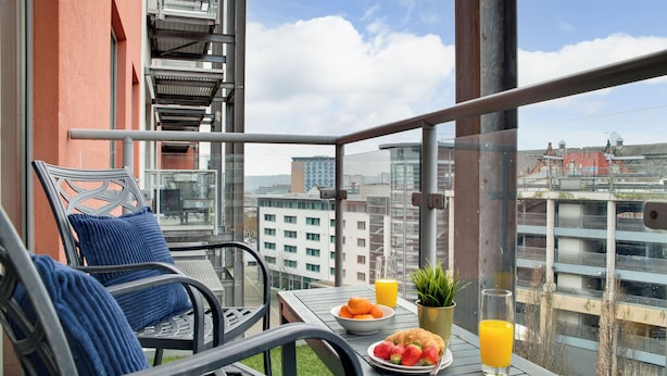 A balcony with a table set for breakfast, including a plate of strawberries, a bowl of oranges, and a glass of orange juice. - Crown Apartments By Week2Week (Newcastle-upon-Tyne)