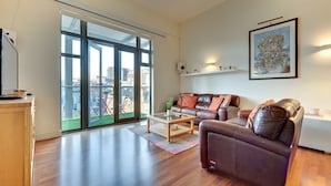 A spacious living room with a large sliding glass door leading to a balcony, a brown leather sofa, a wooden coffee table, and a framed cityscape painting on the wall. - Crown Apartments By Week2Week (Newcastle-upon-Tyne)