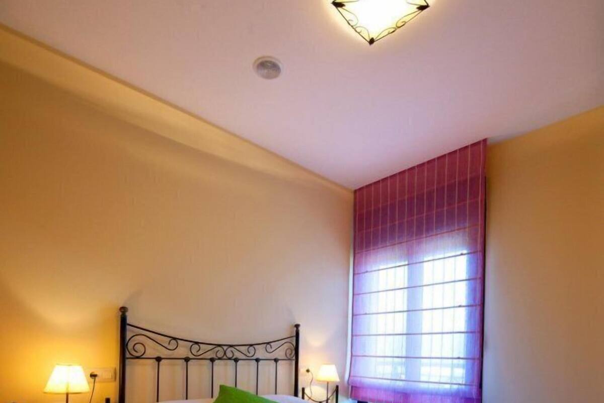 Un dormitorio con cama, lámparas de noche, ventana con cortinas e iluminación en el techo.