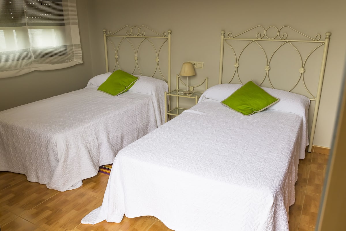 Dos camas individuales con ropa blanca y almohadas verdes, una mesita de noche con lámpara, y una ventana con cortinas traslúcidas.