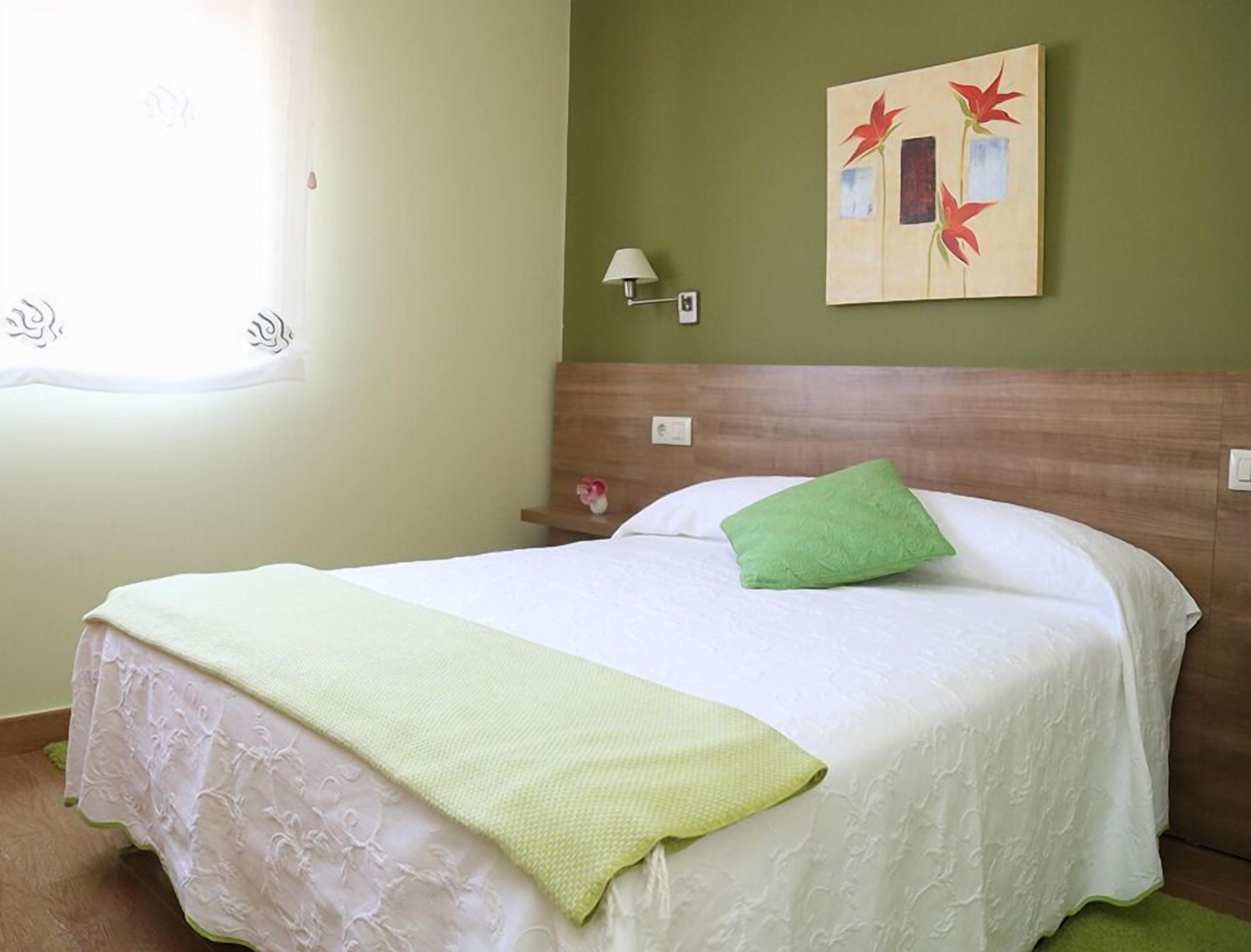 Una cama bien hecha con un cojín verde y una colcha blanca, una lámpara de pared y un cuadro de flores rojas.