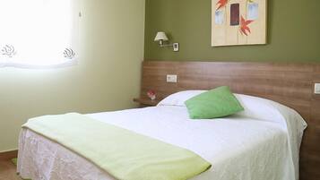 Una cama bien hecha con un cojín verde y una colcha blanca, una lámpara de pared y un cuadro de flores rojas.