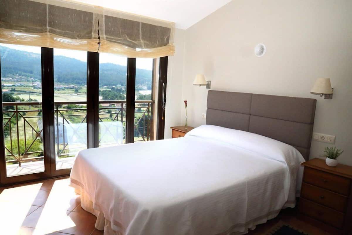 Un dormitorio con una cama grande, dos lámparas de noche, una mesita de noche y vistas a un balcón con un paisaje pintoresco.