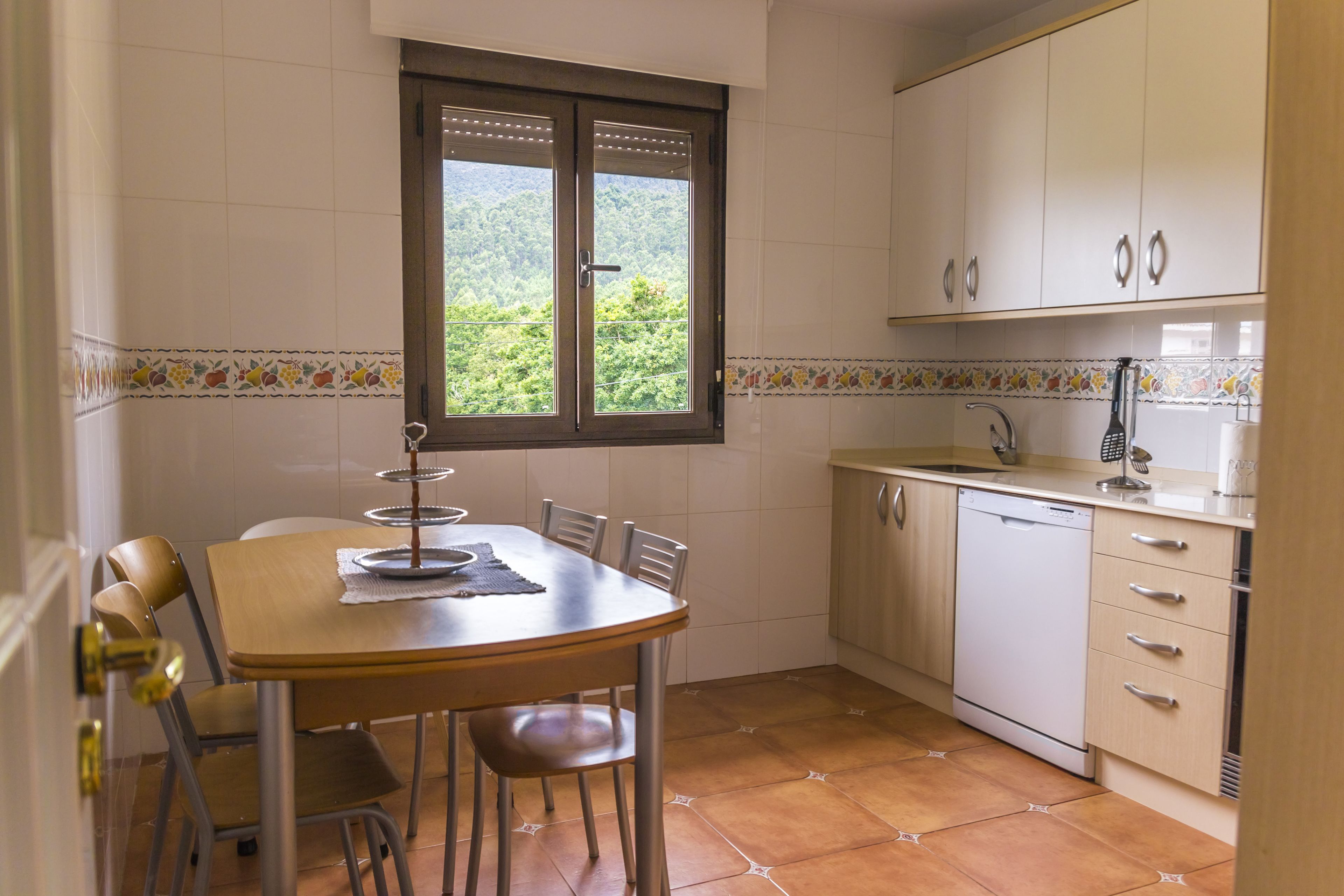 Una cocina con una mesa, sillas, armarios, un ventanal y un lavavajillas.