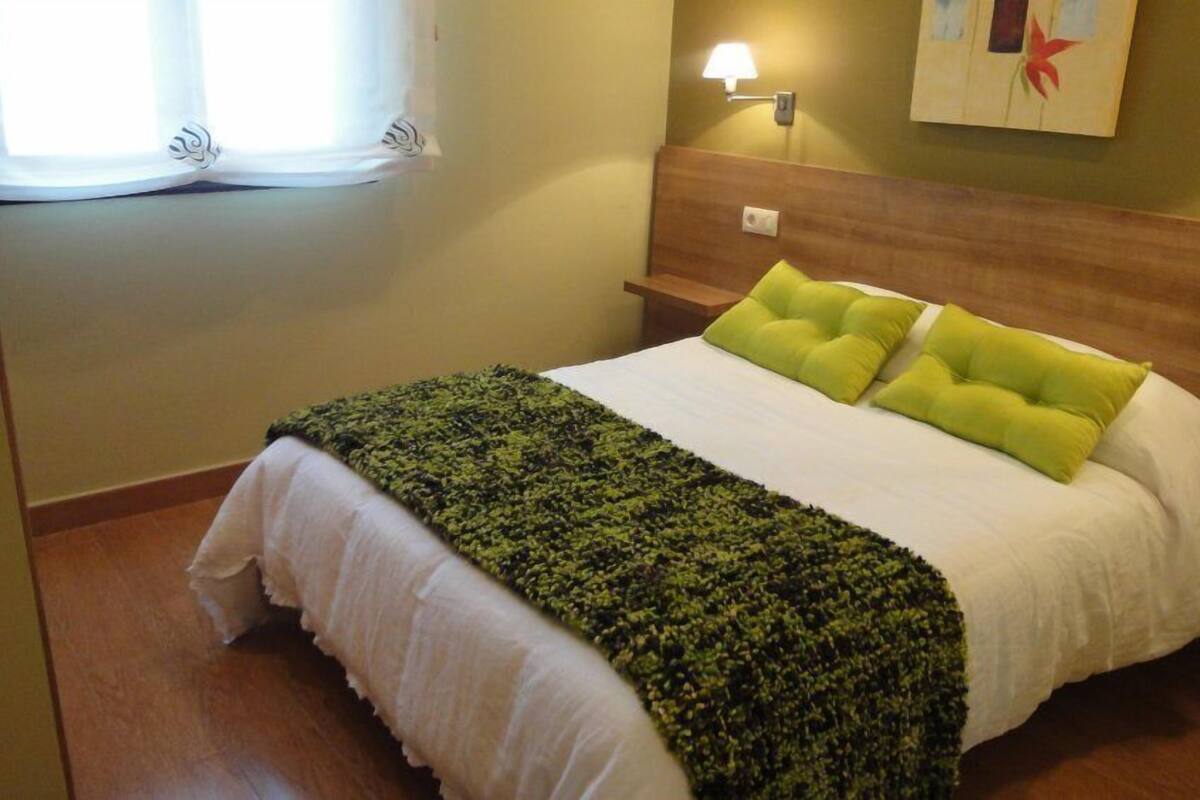 Una cama bien hecha con edredón blanco y almohadas verdes, una manta verde y un cabecero de madera, una lámpara de noche y una ventana con cortinas.
