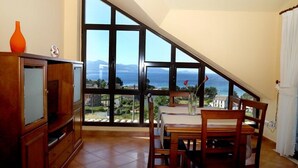 A dining area with a table and chairs, large windows with a view of the sea and mountains, and a wooden cabinet with a vase. - Apartamentos Rurales Abelleira (Muros)