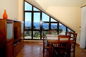 Un comedor con una mesa y sillas, ventanas amplias con vista al mar y a las montañas, y un armario de madera con un jarrón.