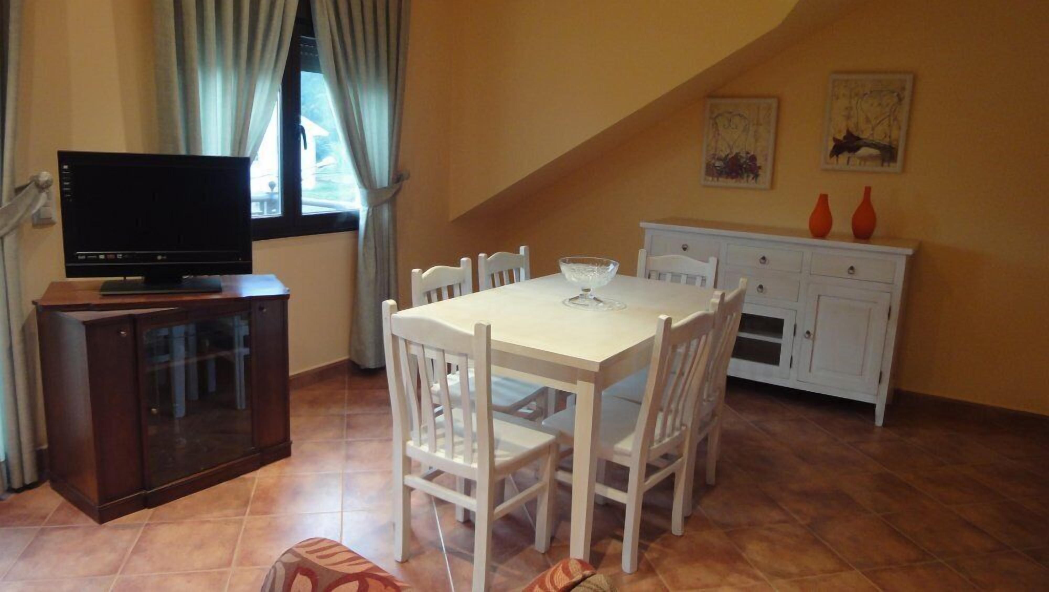 Un coin repas avec une table et des chaises, un téléviseur posé sur un meuble, et un espace cuisine avec des placards et un vase.