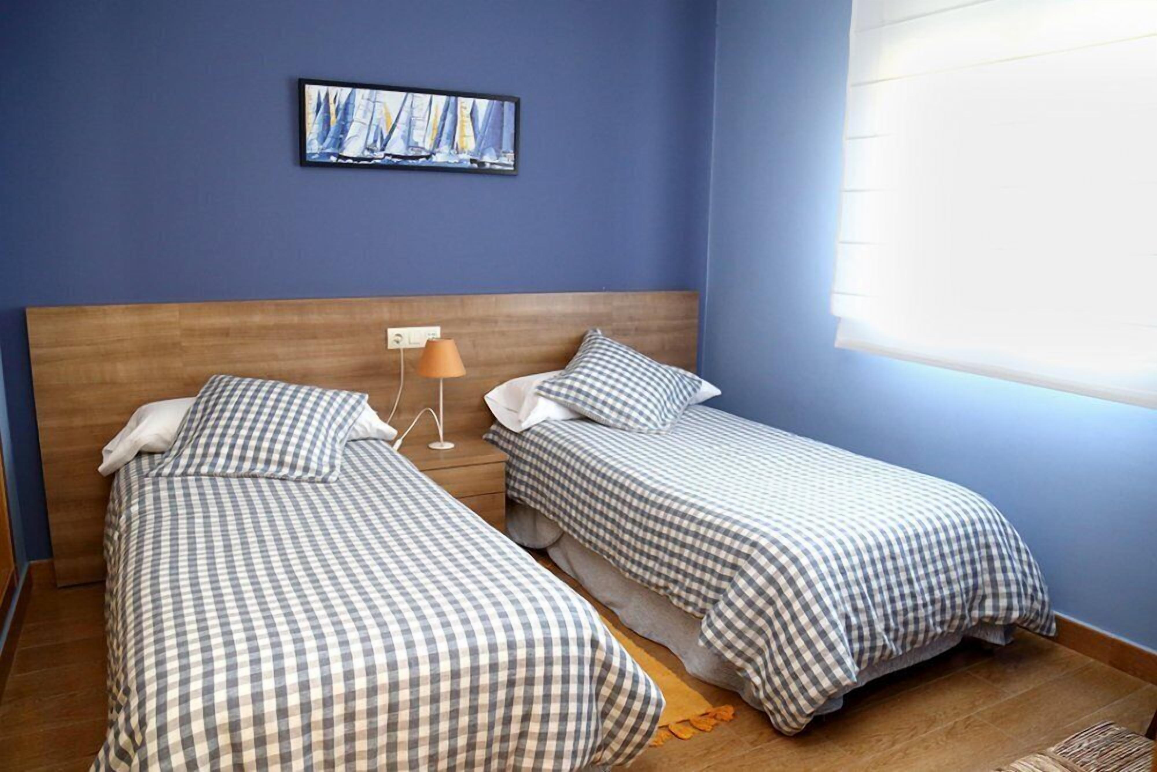 Una habitación con dos camas, cada una con una colcha a cuadros, una mesita de noche con una lámpara y un cuadro en la pared.