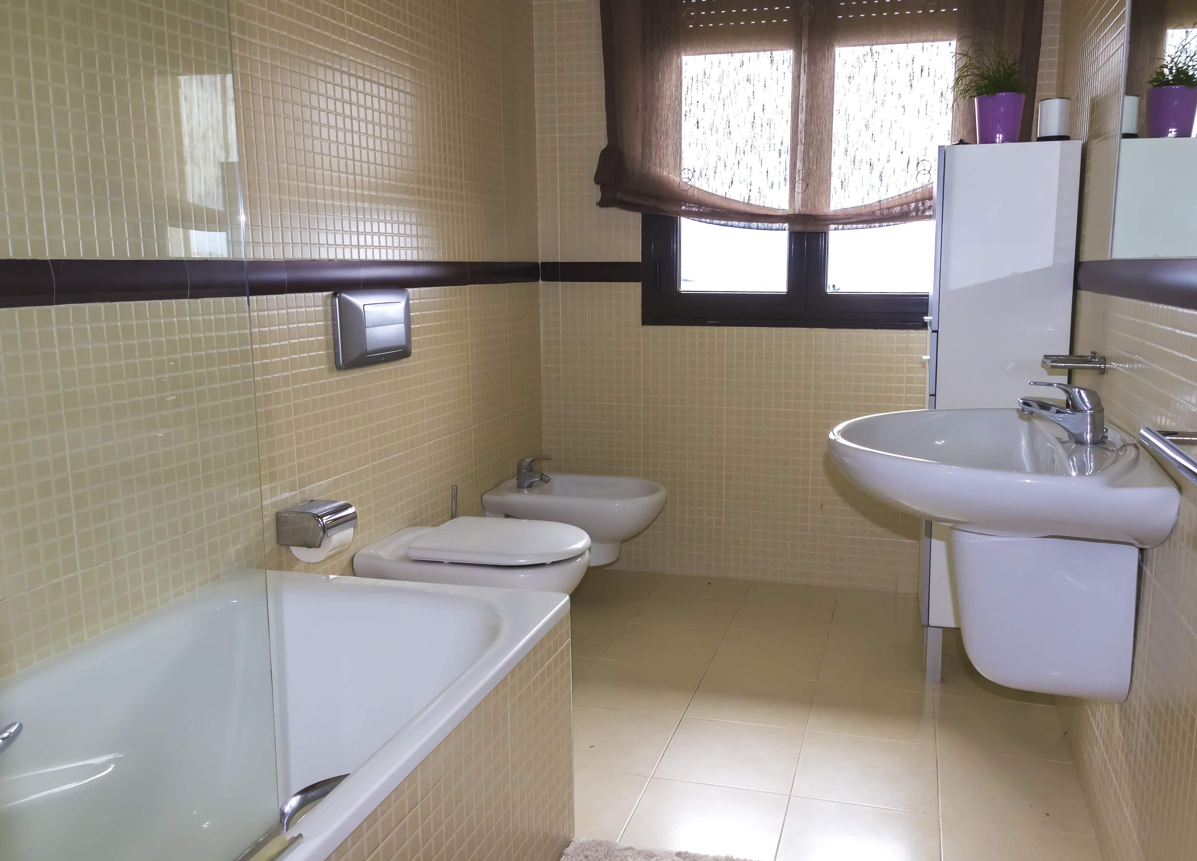 A bathroom with a bathtub, two toilets, a sink, and a window with curtains.