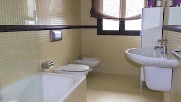 A bathroom with a bathtub, two toilets, a sink, and a window with curtains.