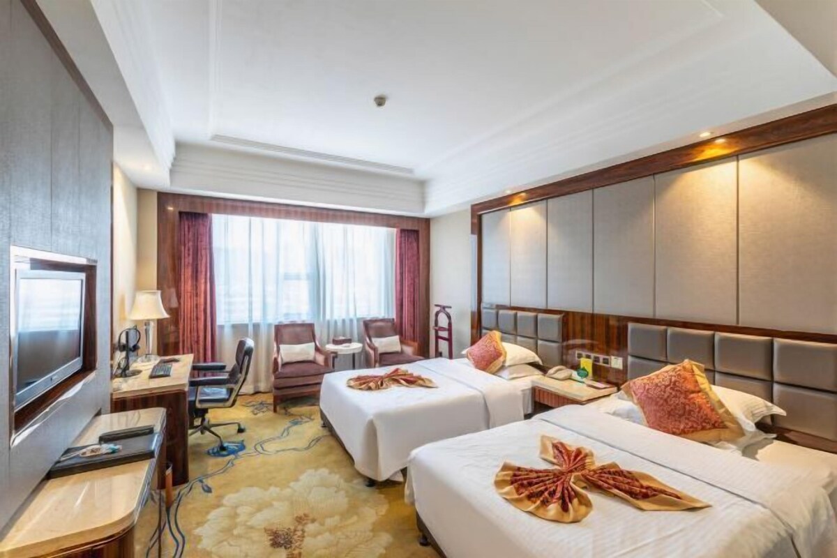 A hotel room with two beds, a desk, a TV, and a large window with curtains.