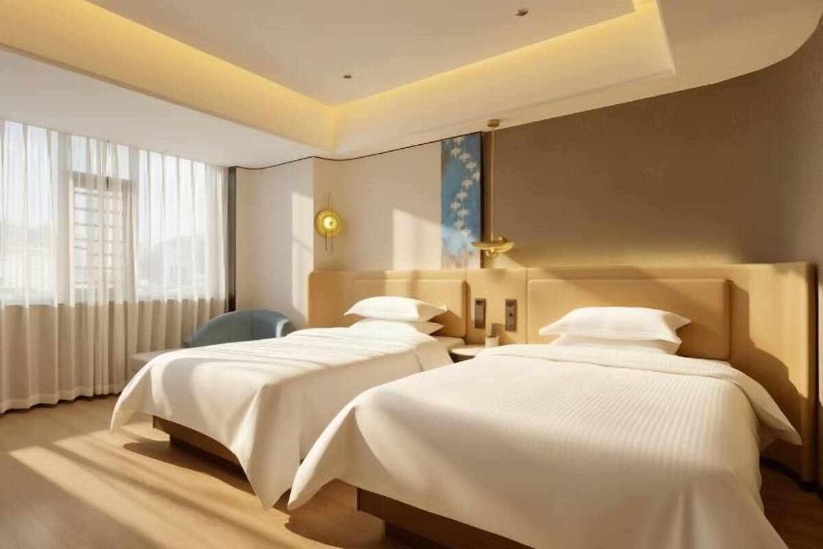 Une chambre d’hôtel avec deux lits, une grande fenêtre avec des rideaux légers, un luminaire fixé au mur et un panneau décoratif.