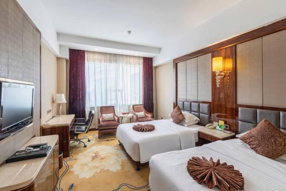 Une chambre d’hôtel avec deux lits, un bureau avec un ordinateur, une télévision, une fenêtre avec des rideaux et une lampe.