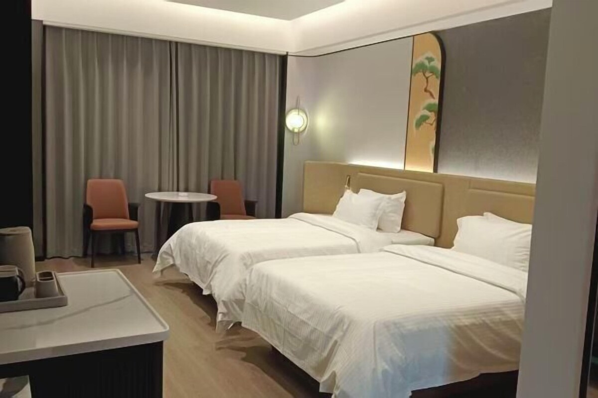 A hotel room with two beds, a small table, a chair, and a large window with curtains.