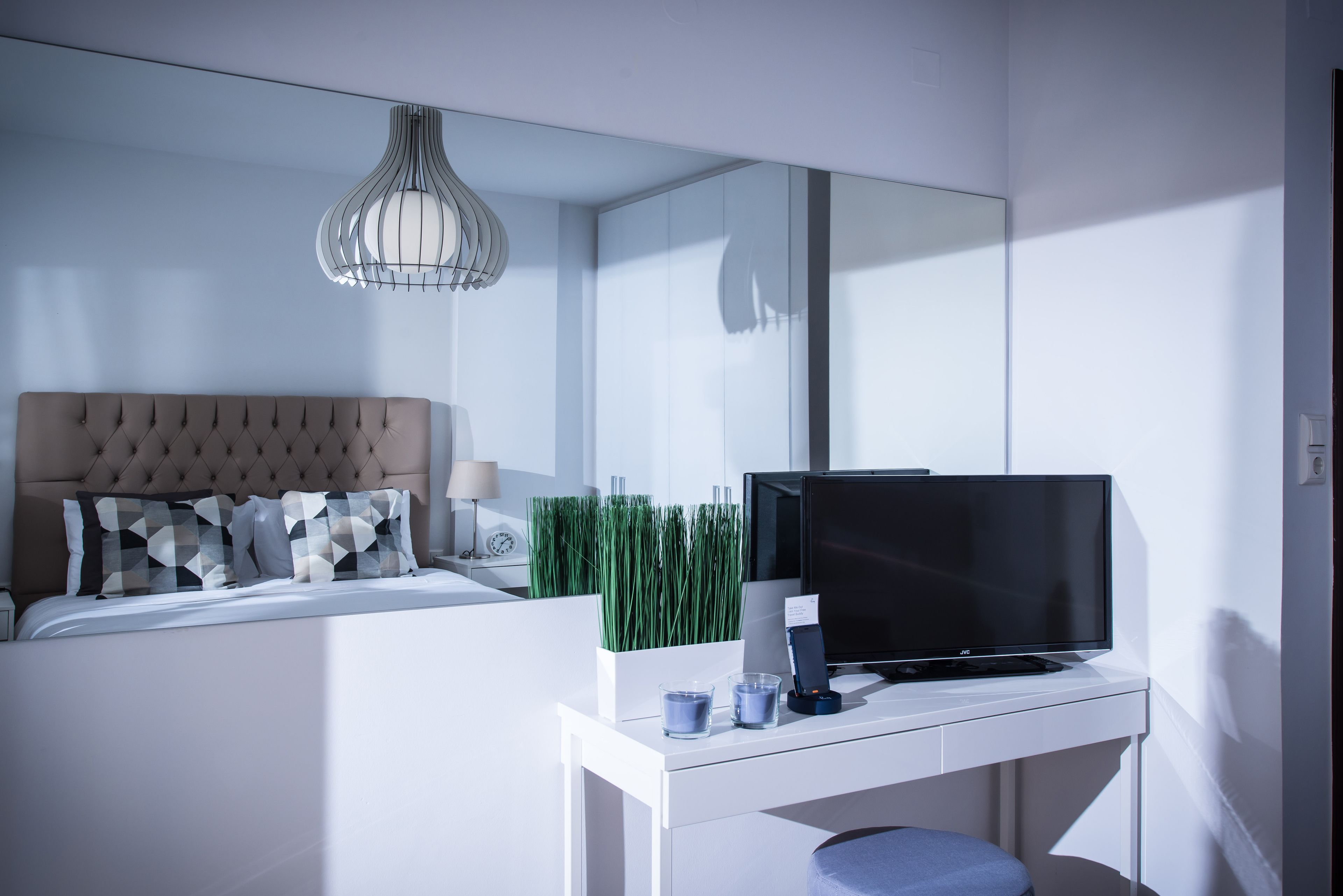 A modern bedroom with a tufted headboard, a flat-screen TV on a white console, a bedside table with a lamp, and a large mirror.