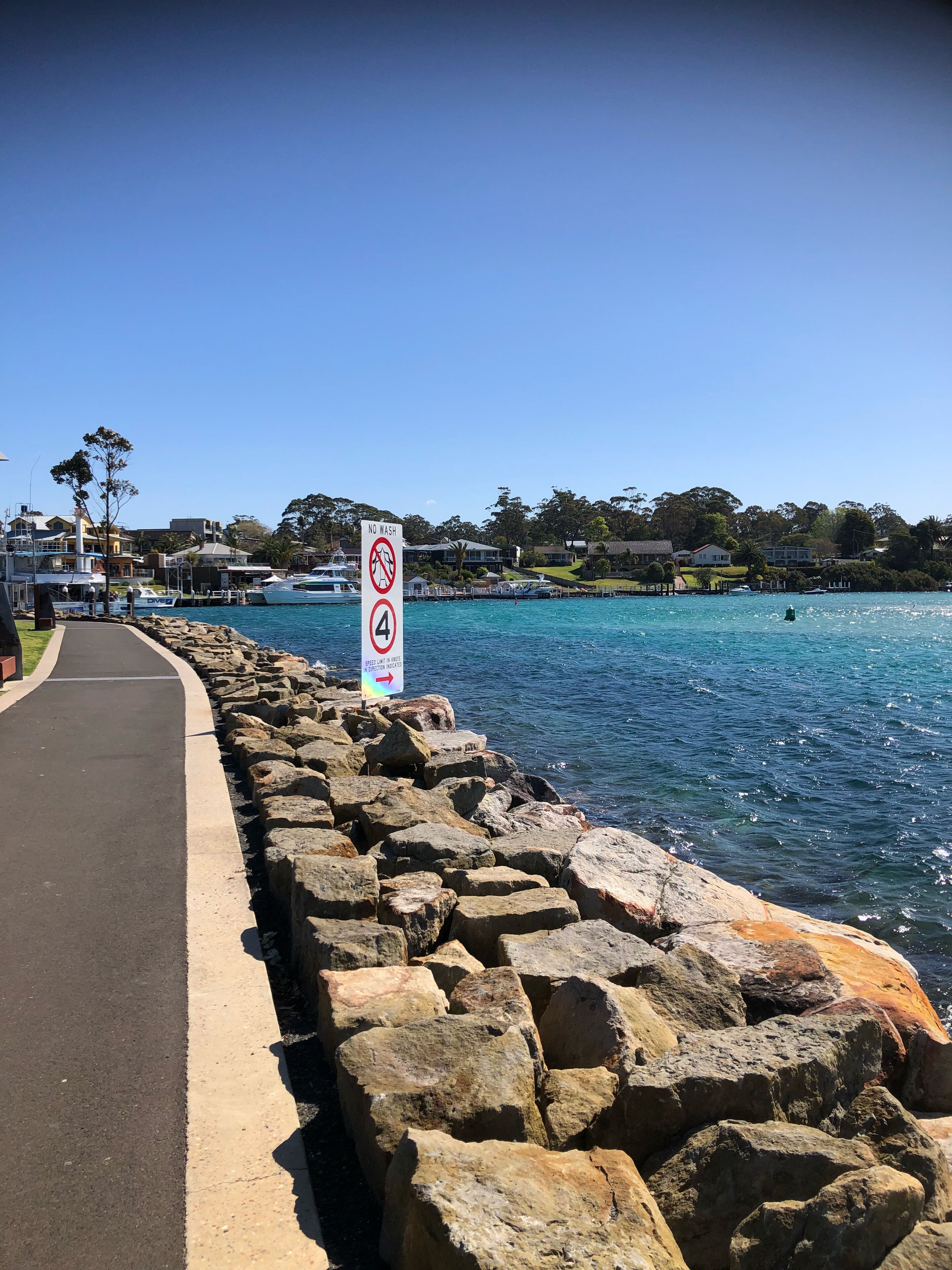 A coastal walkway with a 'No Wash' sign, a speed limit of 4, and a clear view of the sea and houses in the background.