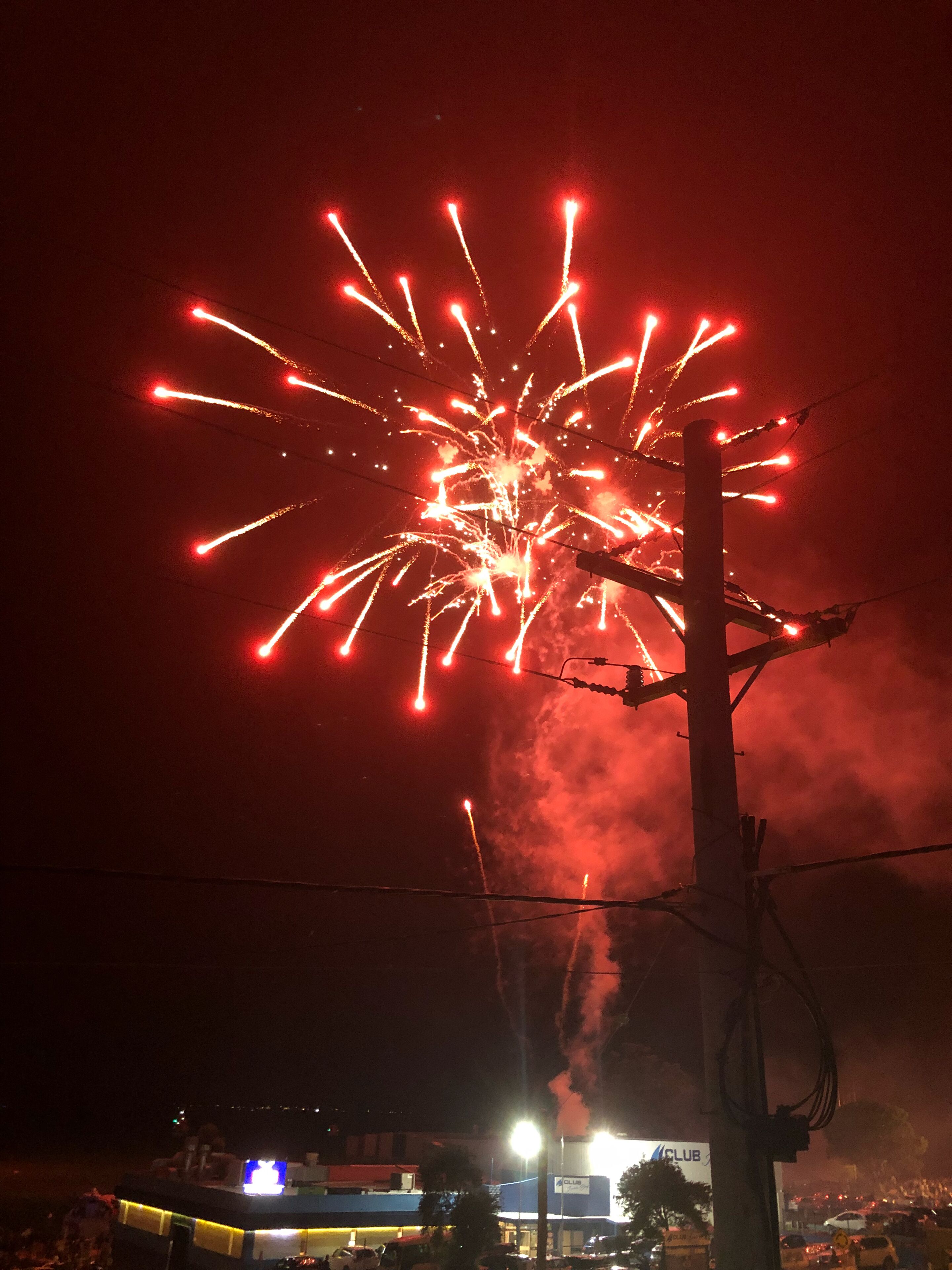 Une scène nocturne avec des feux d’artifice dans le ciel, un bâtiment dont les enseignes sont illuminées et une rue bordée de voitures garées.