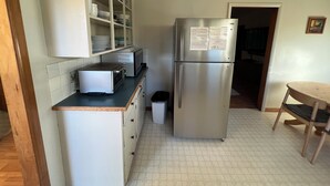 Una cucina compatta con forno a microonde integrato, tostapane e frigorifero in acciaio inox. Si nota un piccolo cestino e una sedia di legno nell'angolo.
