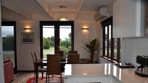 A modern dining area with a large table, chairs, and a red patterned rug. There is a kitchen area with a coffee machine and a window with a view of greenery. - Yolla Beach House A Quiet Relaxing Getaway (Bruny Island)