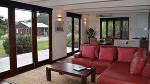 A living room with a red sofa, a coffee table, and a view of a house through large sliding glass doors. - Yolla Beach House A Quiet Relaxing Getaway (Bruny Island)