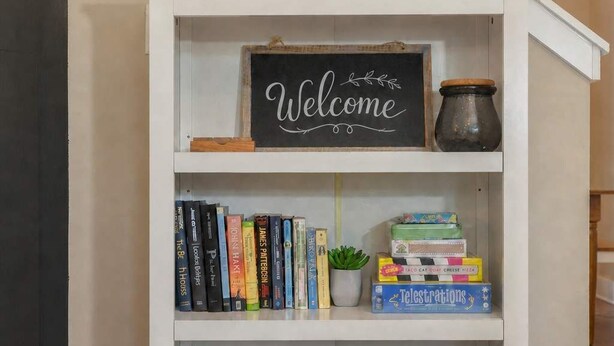 Une bibliothèque blanche ornée d’une pancarte « Bienvenue », avec des livres, une petite plante et un panier en osier.