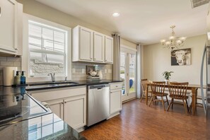 A kitchen with white cabinets, a dishwasher, a window with blinds, a dining area with wooden chairs, and a chandelier. - Pet-Friendly Home: King Bed, 10 Min to Biltmore (Candler)