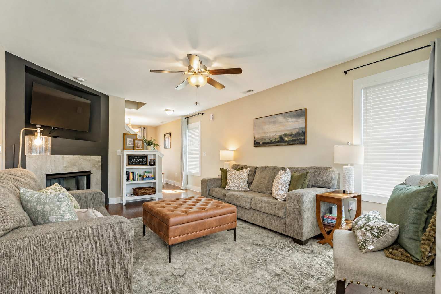 A living room with a fireplace, a ceiling fan, a bookshelf, and a large window with blinds.
