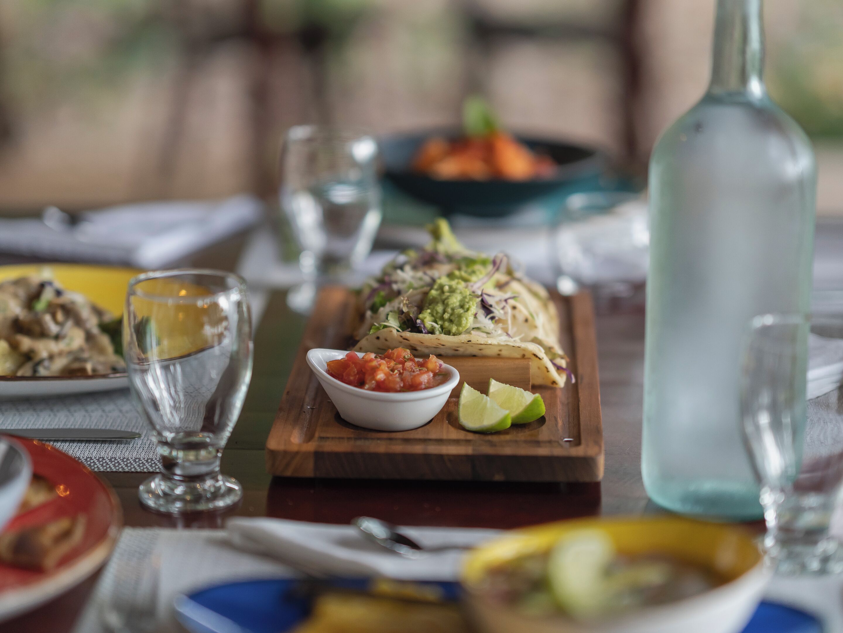 A dining table with a wooden board holding a taco, a small bowl of salsa, and a lime wedge. There are also glasses and a bottle on the table.