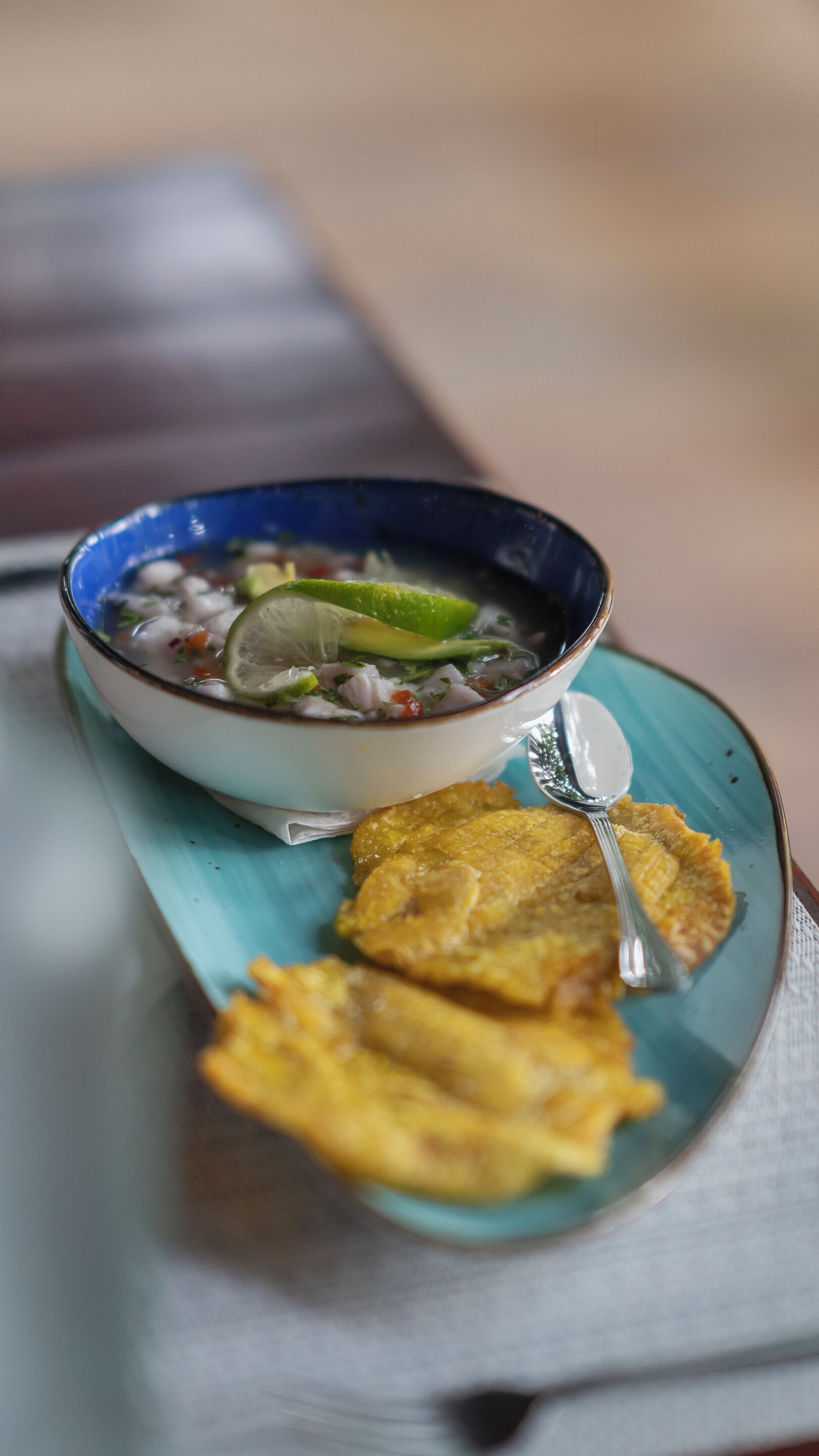 Un bol de soupe, accompagné d’un quartier de citron vert et d’une cuillère, est posé sur une assiette turquoise, avec des tranches de bananes plantains frites à côté.