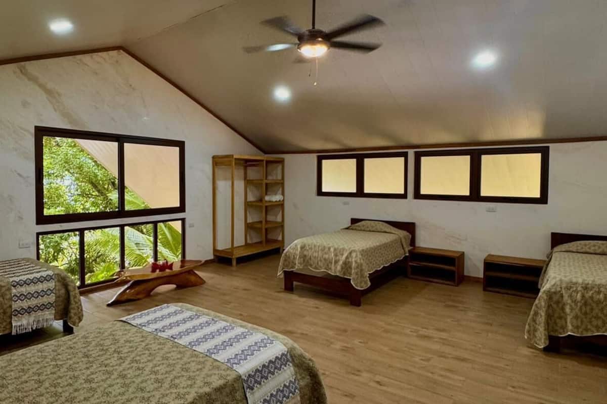 A spacious room with three beds, a ceiling fan, and large windows offering a view of greenery.