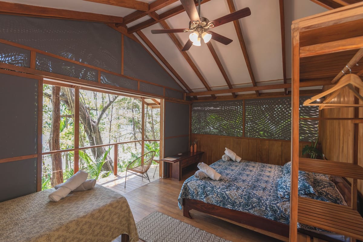 A bedroom with two beds, a ceiling fan, and large windows overlooking a forest.
