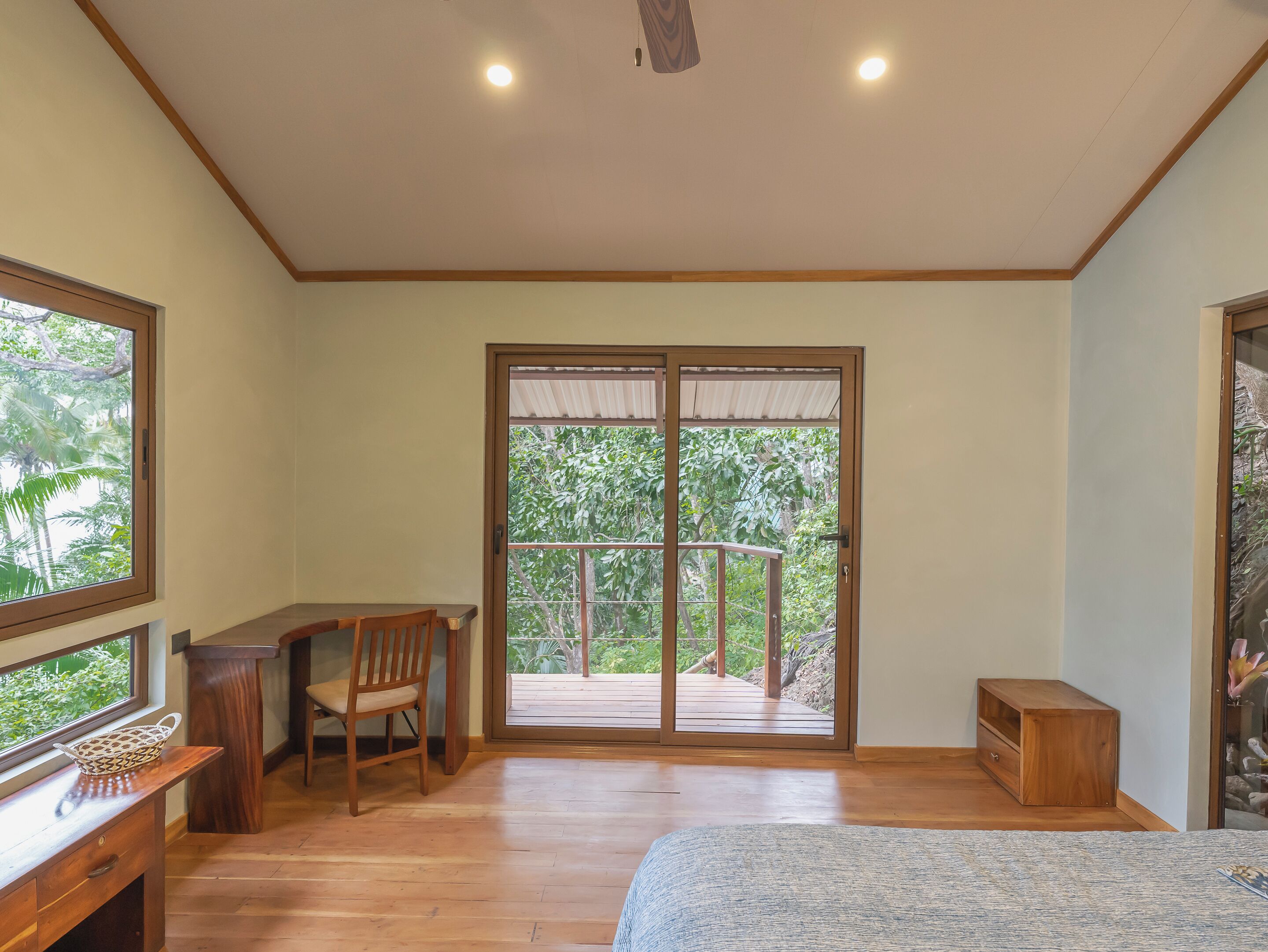 A room with a wooden desk, chair, and a bed. There is a sliding glass door leading to an outdoor deck with a view of greenery.