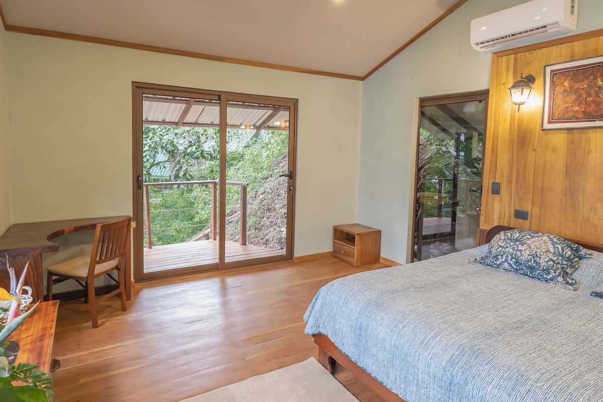 A bedroom with a bed, a desk, and a chair. There is a sliding glass door leading to a balcony with a view of trees.
