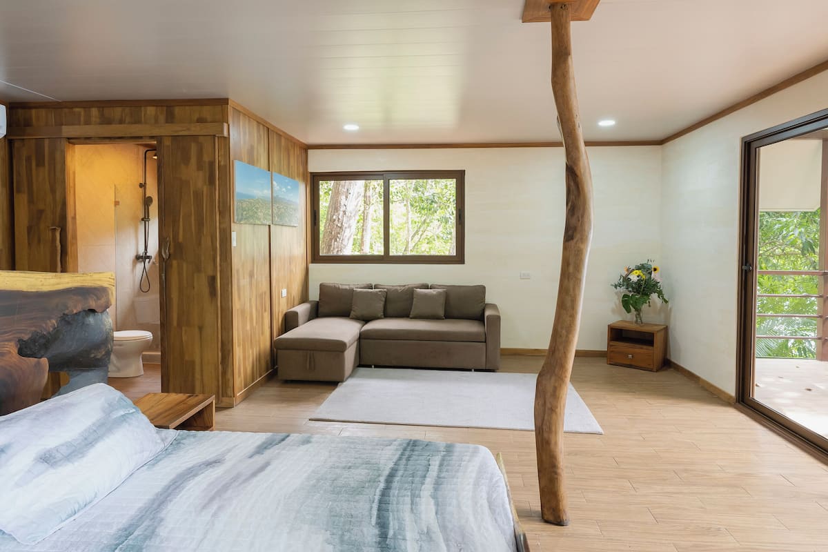 A room with a wooden floor, a sofa, a bed, a wooden pillar, and a window with a view of trees.