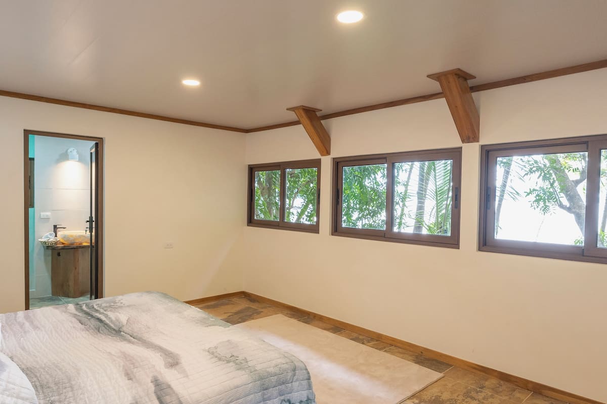 A bedroom with a bed, a small bathroom, and a window with a view of trees.