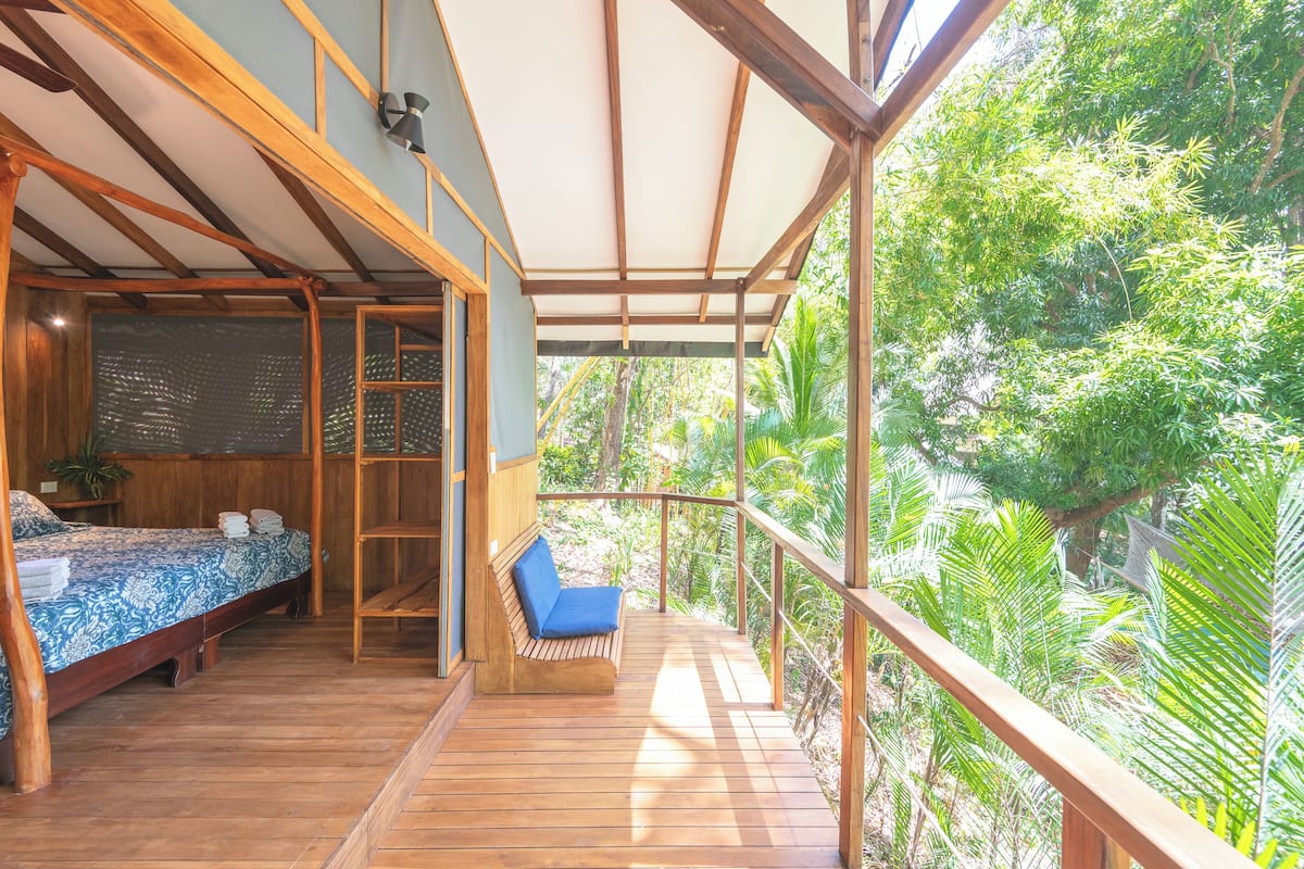 A wooden deck with a bed and a bench, surrounded by lush greenery.