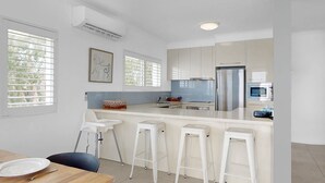 Uma cozinha moderna com armários brancos, uma geladeira de aço inoxidável, micro-ondas e uma área de jantar com mesa de madeira e banquetas.
