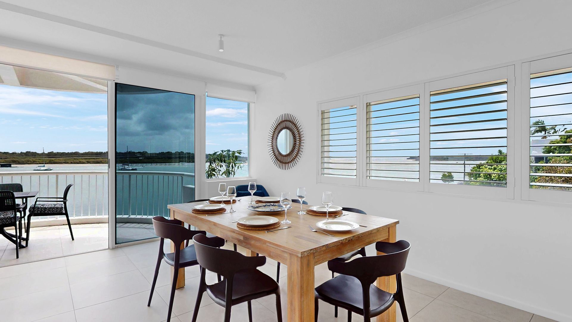 Un comedor con una mesa de madera dispuesta para seis personas, sillas negras, ventanas amplias con persianas horizontales y vista al mar.