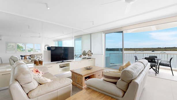 Uma sala de estar moderna com uma televisão grande, assentos confortáveis, uma mesa de centro de madeira e vista para o mar.