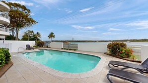 Piscina com vista para a água e barcos, cercada por um muro branco e palmeiras.