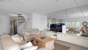 Uma sala de estar moderna com um sofá, mesa de centro e um espelho grande que reflete o ambiente. Uma escada com corrimão é visível ao fundo.