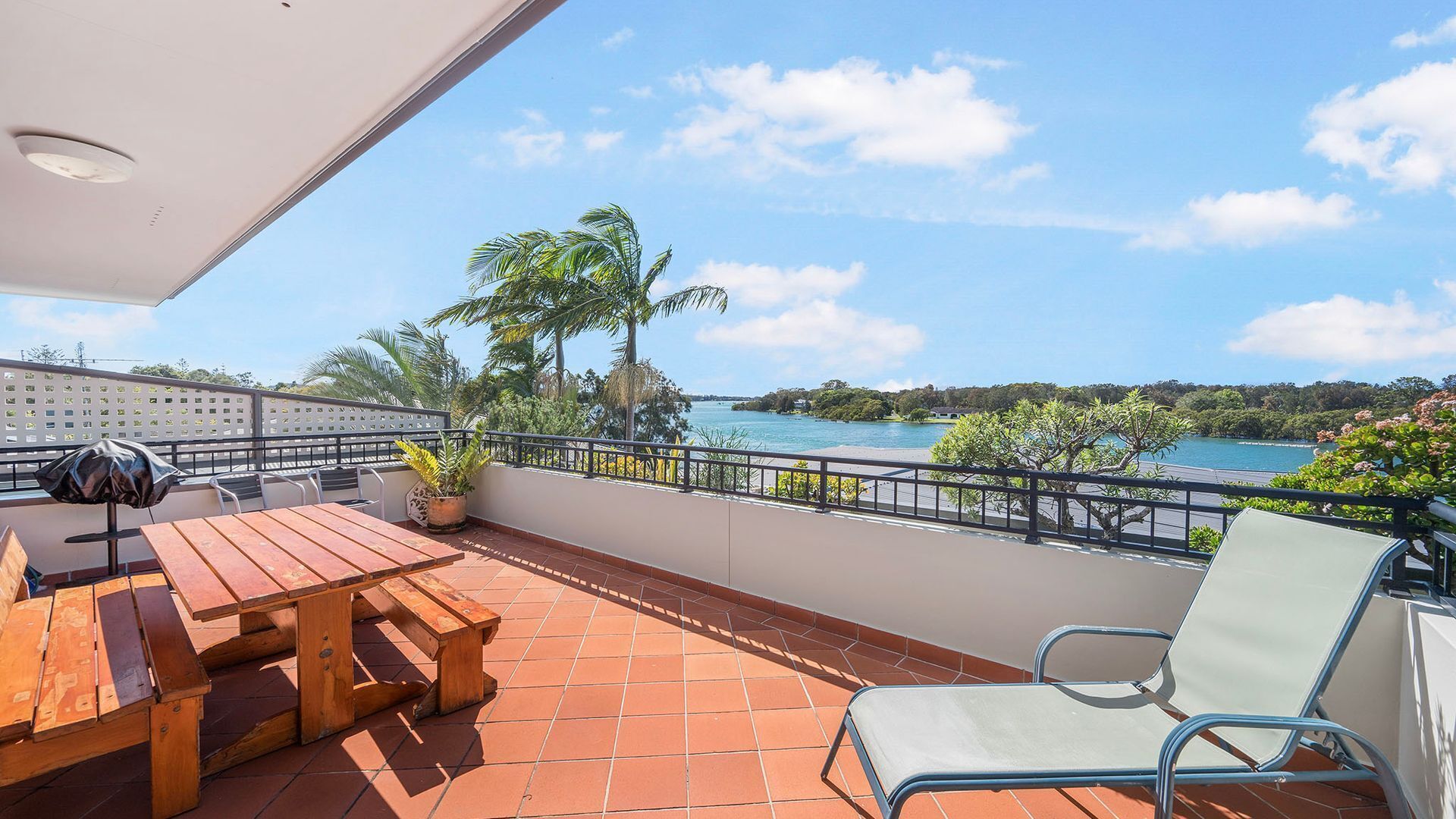 A balcony with a wooden table and chairs, a potted plant, and a view of a body of water and greenery.