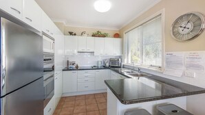 Une cuisine moderne avec des meubles blancs, des appareils en acier inoxydable, un plan de travail en granit noir et une grande fenêtre avec des stores.