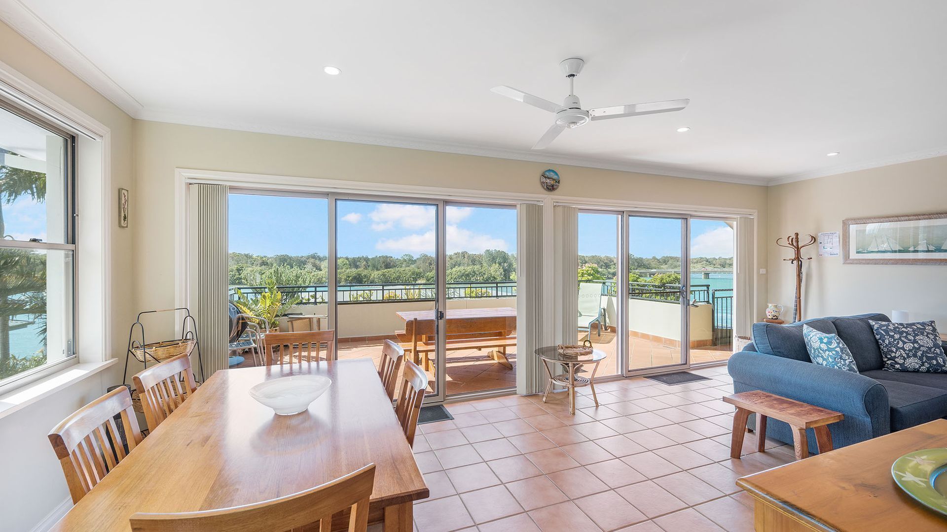 A spacious living area with a dining table, chairs, a sofa, and a ceiling fan. Large sliding glass doors lead to an outdoor balcony with a view of the greenery and water.