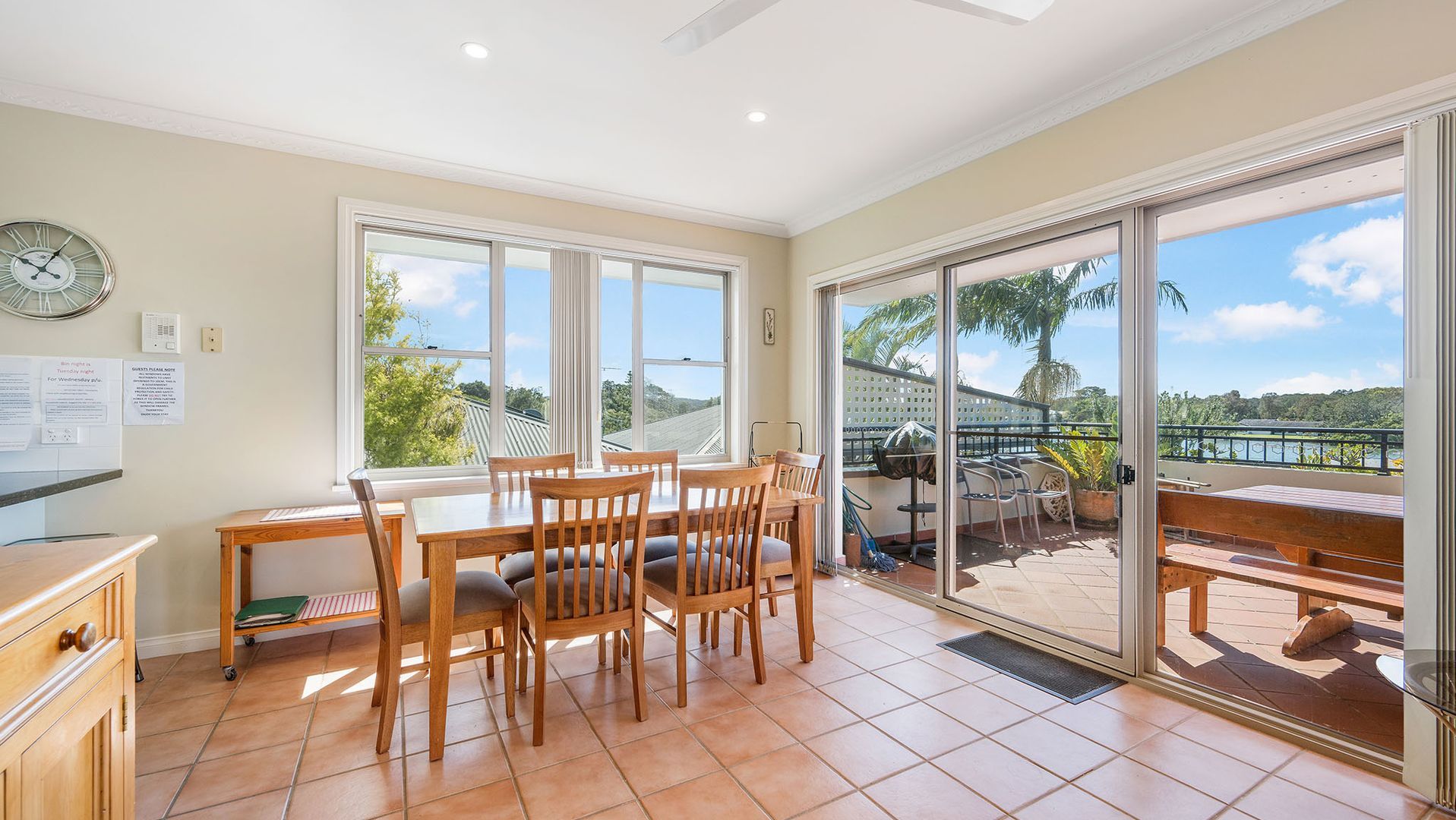 Un coin repas avec une table et des chaises, une horloge au mur et une vue sur un balcon aménagé avec des sièges d’extérieur.