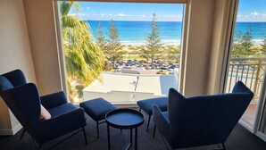 A balcony with two dark blue armchairs and a small round table, overlooking a beach with parked cars and palm trees.