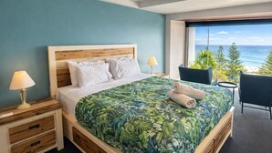 A modern bedroom with a large bed, two bedside tables, a chair, and a view of the ocean.