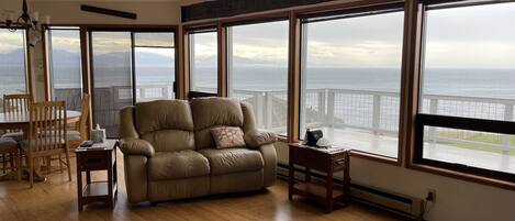 A spacious living room with a large leather sofa, wooden dining table, and chairs. It features large windows with a view of the ocean and mountains.