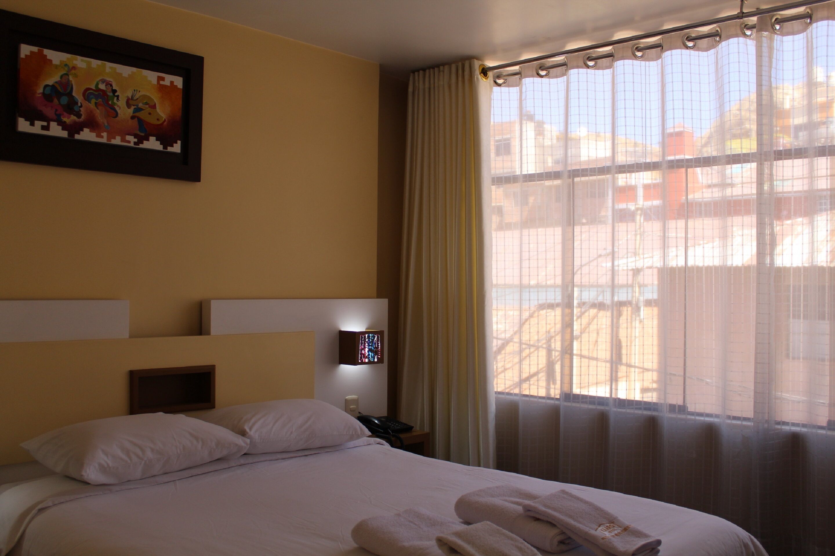 Ein Hotelzimmer mit Bett, Kissen, Nachttisch, Lampe, einem Fenster mit Vorhängen und einem Bilderrahmen an der Wand.